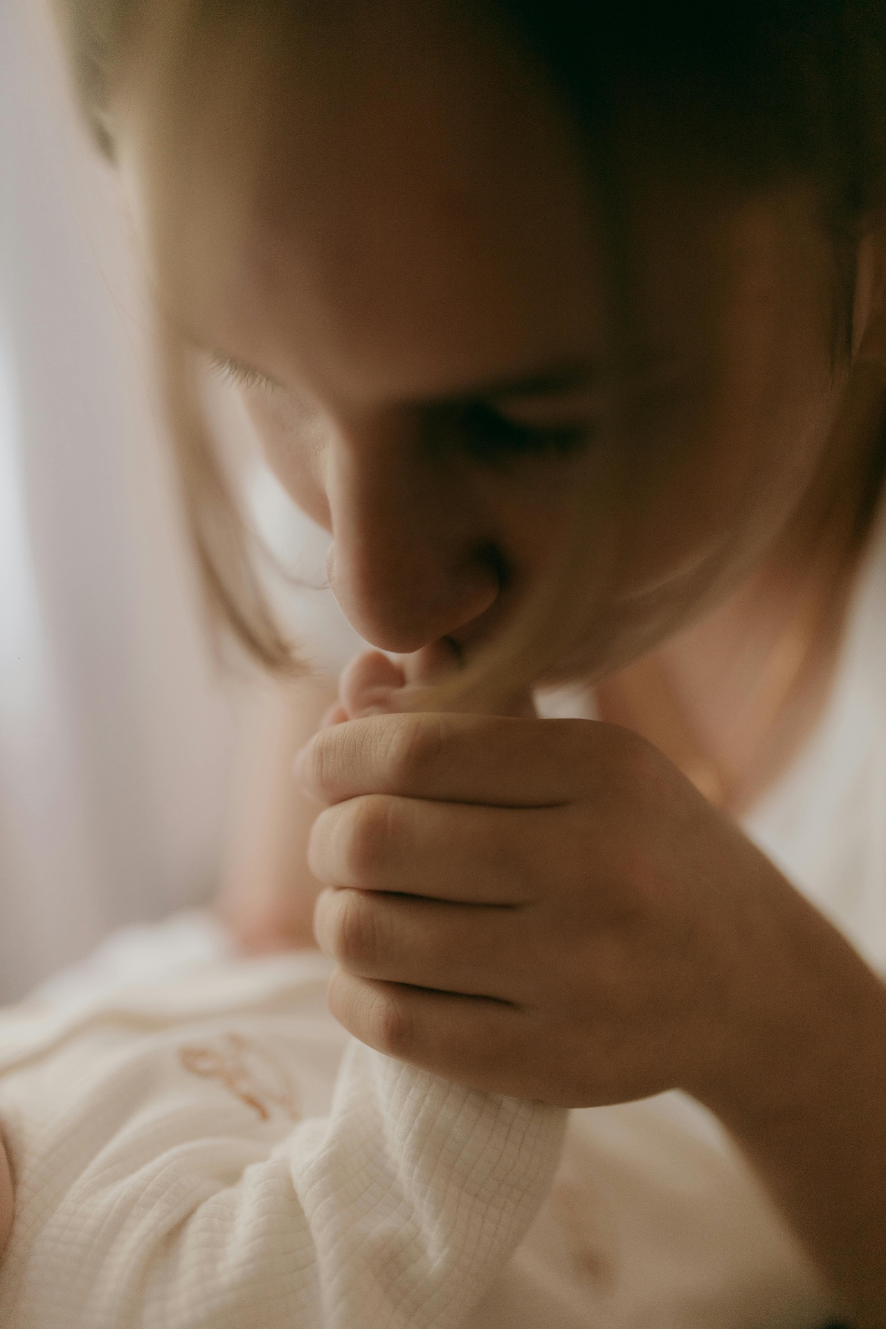 A mother resting on a bed in soft daylight, her newborn's small hand held inside her own.